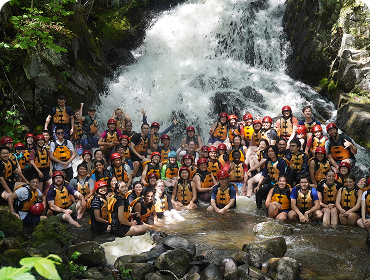 A large group of kids in rafting gear posing in front of a waterfall